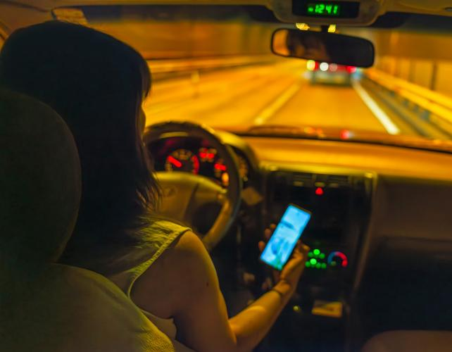 una foto scattata sopra la spalla di una giovane donna alla guida che tiene il telefono mentre guida di notte.