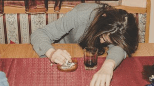 Una donna che sembra ubriaca, con una bevanda e una sigaretta nei paraggi.