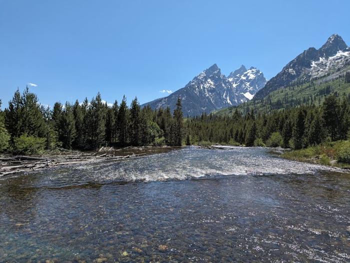 Un ampio e tranquillo fiume scorre in una fitta foresta. Imponenti picchi frastagliati perforano il cielo. La neve è visibile nelle crepe della montagna.