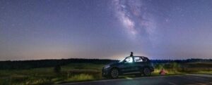 un'auto parcheggiata sul lato della strada in un campo remoto. la via lattea e un meteorite sono visibili nel cielo notturno. una persona è seduta sopra l'auto, un'altra persona è seduta accanto a essa, entrambe guardando il cielo.