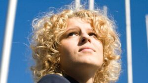 Giovane adulto con capelli biondi ricci, che guarda in alto con un cielo luminoso sullo sfondo