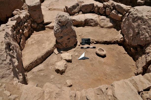 Una pietra oblunghe in piedi nel mezzo di un recinto circolare di pietre con pareti e senza tetto. Un mortaio si trova a destra della pietra centrale, e marcatori archeologici sono stati posti alla base della pietra.