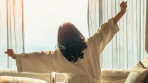 Una persona con lunghi capelli scuri che guarda una mattina soleggiata da una finestra, allungando entrambe le braccia in aria