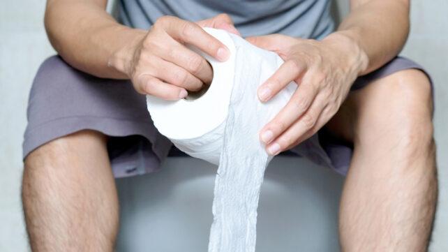 Un uomo seduto su un water, completamente vestito, che tiene un rotolo di carta igienica. Per illustrare una connessione tra la frequenza dei movimenti intestinali e la salute generale