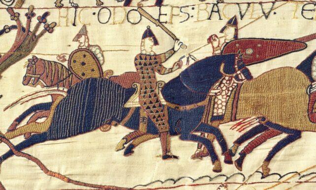 una sezione della tappezzeria di Bayeux che mostra una scena di battaglia ricamata in giallo, blu e rosso, su uno sfondo di stoffa chiara
