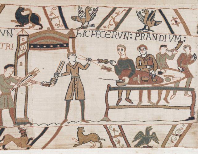 una scena nella tappezzeria di Bayeux che mostra tre soldati che banchettano allegramente a un tavolo. a sinistra, un servitore offre carne arrosto su lunghi bastoni. a destra, un soldato suona un corno.