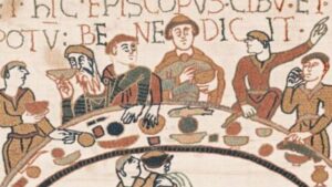 una sezione della tappezzeria di Bayeux che rappresenta sei uomini attorno a un tavolo a forma di mezzaluna che mangiano e bevono. un uomo in basso serve vino.