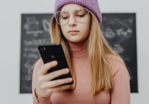 ragazza con occhiali e berretto viola che guarda il suo smartphone con una lavagna sullo sfondo