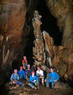 Un gruppo di 7 speleologi in tute blu e bianche e caschi rossi posa davanti a una stalagmite alta 8 metri, decorata in modo intricato, in una caverna buia.