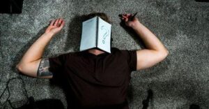 uomo che dorme sul pavimento con il viso coperto da un quaderno aperto, con un grande punto interrogativo scritto su una delle pagine