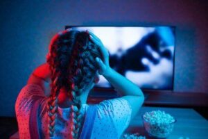 giovane donna vista di spalle che tiene le mani sulla testa mentre guarda un film in TV, l'illuminazione è scura con rosso e viola. C'è una ciotola di popcorn sul tavolo di fronte a lei. Ha due trecce nei capelli e indossa una salopette e una maglietta a pois.