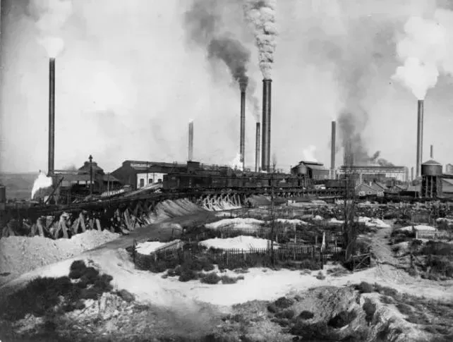 una foto in bianco e nero di un impianto industriale di fusione, con camini che rilasciano fumi nell'aria.