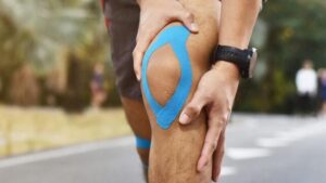 Le mani di un uomo che tiene il ginocchio mentre è in pausa dalla corsa all'aperto. Il suo ginocchio è fasciato con del nastro sportivo blu
