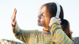 una ragazza adolescente bianca con i capelli castani raccolti in una coda di cavallo indossa cuffie over-ear e tiene la mano alzata, guardando il retro di essa. è all'aperto, di fronte a dei pannelli solari e a una recinzione a catena.