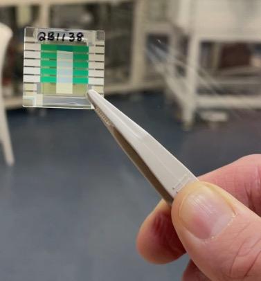 Immagine ravvicinata di una mano che utilizza delle pinzette metalliche per tenere un oggetto sottile, quadrato, verde e bianco.