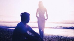 Silhouette di un uomo seduto sulla riva del mare mentre guarda una donna in abito bianco che si avvicina a lui. L'immagine sembra catturare un ricordo lontano