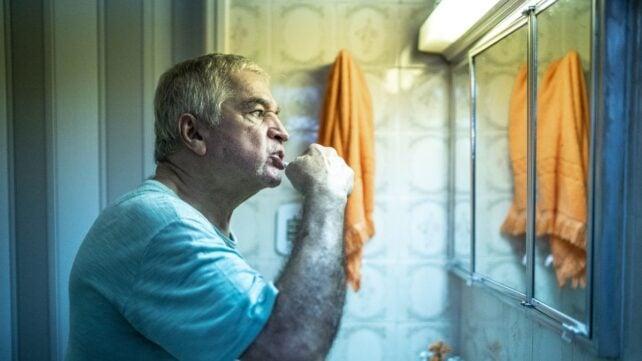 Uomo con capelli grigi che si lava i denti davanti allo specchio del bagno, con scarsa illuminazione e con un asciugamano arancione appeso al muro.
