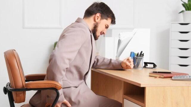 Un uomo è seduto a una scrivania di lavoro tenendosi l'anca e facendo una smorfia come se avesse dolore