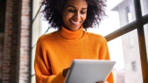 Donna afroamericana sorridente in un luminoso maglione arancione e con un tablet o laptop al lavoro