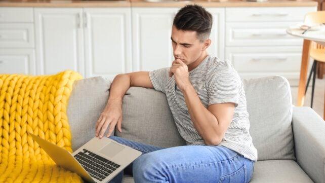 Un giovane uomo bruno seduto su un divano grigio, che guarda un laptop aperto