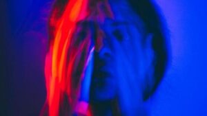 Un intenso ritratto astratto di una persona che si copre il viso con le mani, catturato con esposizione prolungata e luci al neon rosse e blu vibranti e sfocate