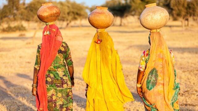 Tre donne in abiti colorati portano grandi recipienti d'acqua sulla testa attraverso un paesaggio rurale secco nel Rajasthan, India.