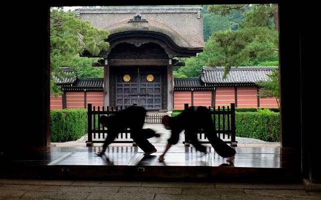 Due monaci lucidano un sentiero di legno del tempio, silhouettati contro un tradizionale cancello e giardino di un tempio giapponese.