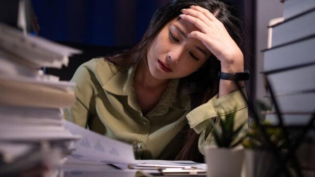 Una donna in una maglietta verde che sembra stanca, con molto lavoro sulla sua scrivania