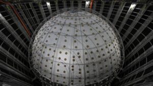 Grande rivelatore di neutrini JUNO sferico all'interno di una camera sotterranea, circondato da cavi, supporti metallici e pannelli murali scuri.