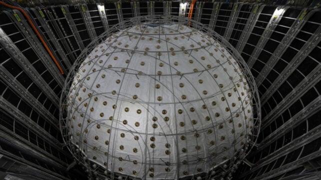 Grande rivelatore di neutrini JUNO sferico all'interno di una camera sotterranea, circondato da cavi, supporti metallici e pannelli murali scuri.