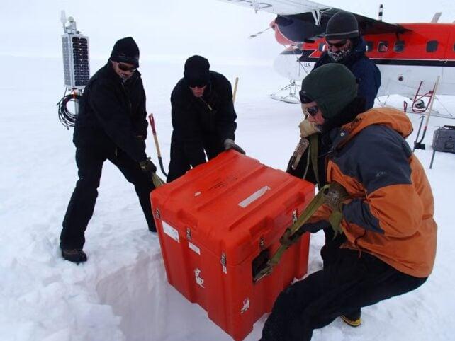 I ricercatori in abbigliamento per il freddo scaricano una grande valigia arancione da un aereo su un sito di ricerca innevato in Antartide.