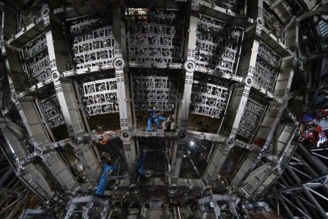 Lavoratori in tute protettive blu installano componenti all'interno del grande rivelatore sferico JUNO, circondato da telai metallici e supporti per sensori circolari.