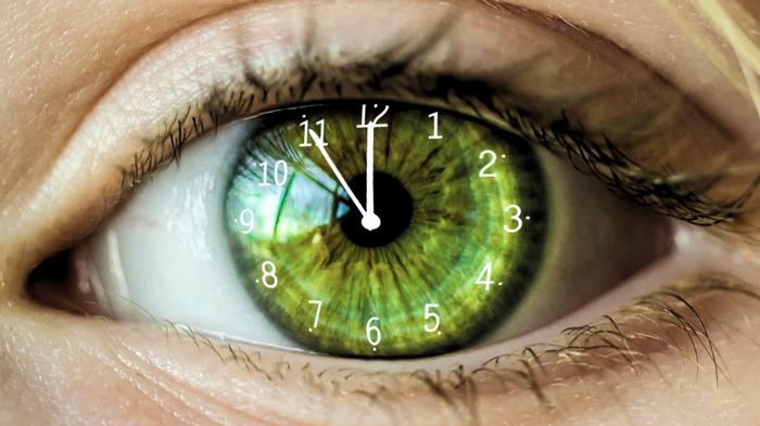 Primo piano di un occhio umano verde con numeri e lancette dell'orologio sovrapposti all'iride, simboleggiando l'invecchiamento biologico e il passaggio del tempo.