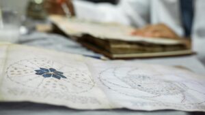 una persona lavora a un manoscritto sullo sfondo, mentre in primo piano si vede una pagina di pergamena del manoscritto Voynich