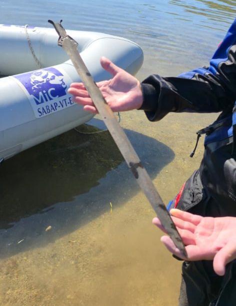 una persona dalla pelle chiara indossa una muta da sub e tiene in mano una spada dell’età del bronzo, mostrandola per tutta la sua lunghezza; sullo sfondo c’è una barca