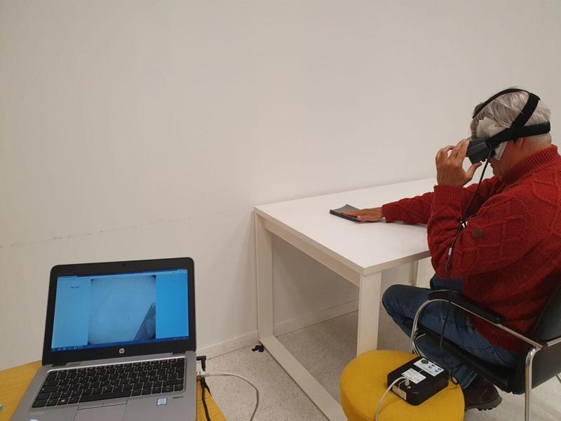 None Un partecipante seduto indossa occhiali speciali e tocca un quaderno su un tavolo bianco; nelle vicinanze si vedono un laptop e apparecchiature collegate.