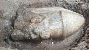 La testa di una statua giace orizzontalmente a terra. Raffigura un uomo che indossa un copricapo a punta.