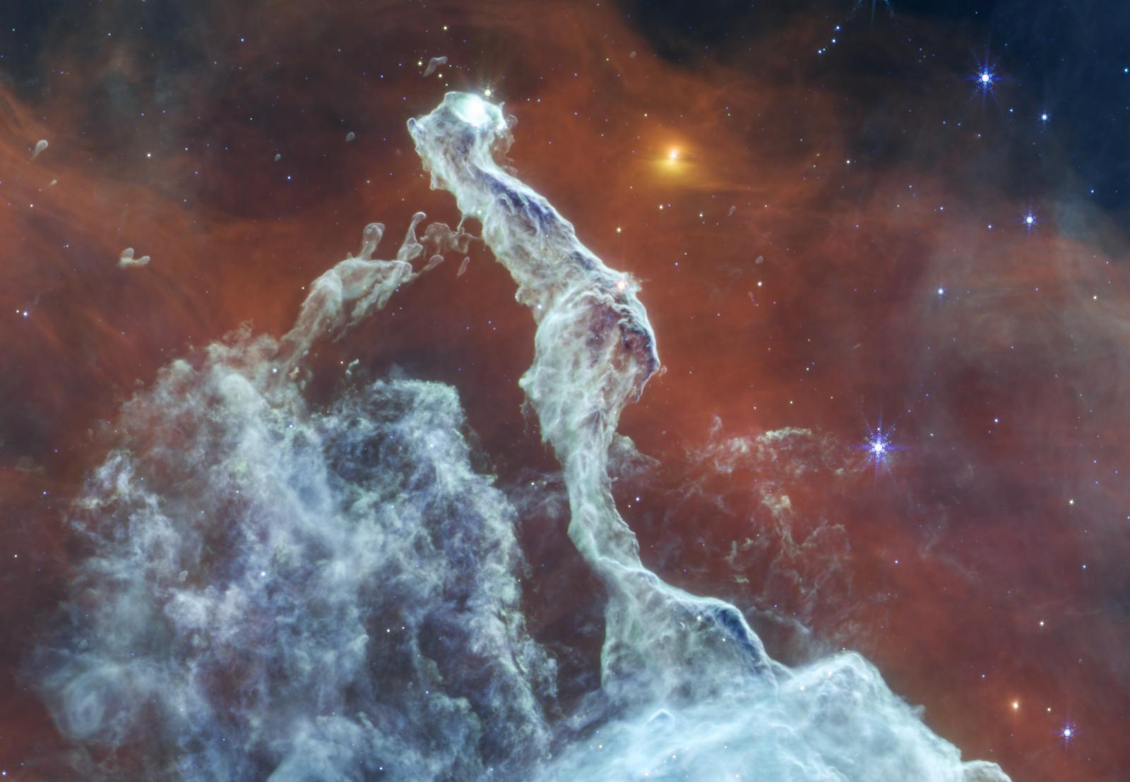 Immagine nel medio infrarosso della Nebulosa Testa di Gatto ripresa dal telescopio spaziale James Webb. Al centro si trova un filamento di polvere grigio-bluastro, che si estende dal bordo inferiore quasi fino a quello superiore. Ha una forma stretta e irregolare, simile a un gatto seduto di spalle rispetto all’osservatore, con la coda rivolta verso il basso. La coda termina nel terzo inferiore dell’immagine e si allarga formando una punta triangolare di polvere. La testa del gatto è girata verso destra e mostra un occhio bianco luminoso. Lo sfondo è perlopiù rosso; negli angoli in alto a sinistra e a destra si intravede lo sfondo nero dello spazio. Alla sinistra del gatto si trova un raggruppamento a forma di cupola di deboli filamenti di polvere. Nell’immagine sono sparse stelle gialle e blu, alcune con le caratteristiche punte di diffrazione a otto punte di Webb.