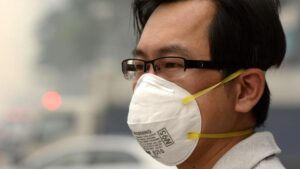 Un uomo indossa occhiali e una mascherina bianca. Sullo sfondo è visibile lo smog.